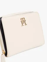 Billetera Tommy Hilfiger color crema con cierre de cremallera y botón a presión. Cuenta con compartimento interior para billetes y múltiples ranuras interiores para tarjetas. Luce el monograma TH metálico en la parte delantera.