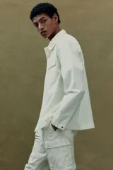 Sobrecamisa color crema con cuello inglés, cierre frontal con botones a presión, dos bolsillos superiores con solapa y puños ajustables con botón a presión.
