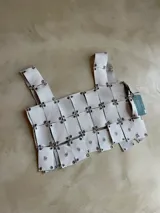 Top blanco de cuero genuino de ovino, compuesto por paneles cuadrados unidos por herrajes plateados. Presenta un diseño bordado a mano con forma de estrella en algunos paneles. Tiene tirantes anchos.