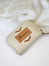 Billetera pequeña de cuero color crudo con cierre metálico y monedero interno. Presenta costuras decorativas en forma de espiga.