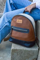 Mochila de cuero color tostado, con base y espalda en negro y tapa superior en blanco. Tiene un bolsillo frontal con cierre y un pequeño aplique de cuero con el logo de la marca.