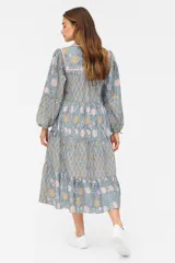 Vestido largo de corte holgado con estampado floral en tonos celeste, rosa y dorado. Tiene cuello redondo con lazo y borlas decorativas, mangas 3/4 con lazos en los puños y falda con volantes.