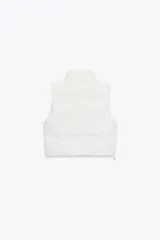 Chaleco blanco acolchado con cuello alto y cierre frontal con cremallera. Tiene bolsillos delanteros con cierre de cremallera oculta y bajo ajustable con cordones elásticos laterales.