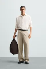 Pantalón de corte holgado (wide fit) en color beige, confeccionado en tejido con mezcla de lyocell. Presenta pliegues frontales en la cintura, bolsillos laterales y bolsillos traseros de doble vivo. Cuenta con cierre frontal de cremallera y botón.