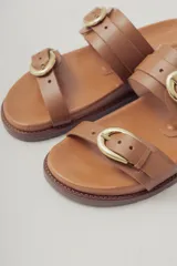 Sandalia plana de cuero color caramelo, estilo 'slide' o 'dos tiras', con hebillas ajustables de metal dorado en cada tira.