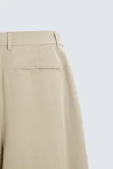 Pantalón de corte holgado (wide fit) en color beige, confeccionado en tejido con mezcla de lyocell. Presenta pliegues frontales en la cintura, bolsillos laterales y bolsillos traseros de doble vivo. Cuenta con cierre frontal de cremallera y botón.