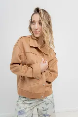 Chaqueta color beige de corte recto, estilo camisa, confeccionada en gamuza con elastano. Presenta cuello camisero, cierre frontal con botones metálicos, bolsillos grandes en el frente y charreteras en los hombros.