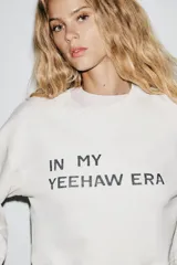 Sudadera corta color blanco hueso, de cuello redondo y manga larga. Tiene un estampado con la frase 'IN MY YEEHAW ERA' en color negro.