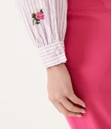 Blusa de algodón a rayas verticales blancas y lilas, con bordado de rosas rosadas. Tiene cuello redondo con volados, mangas largas y volados en los hombros.