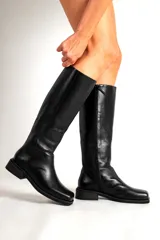 Bota de cuero negra, de caña alta y corte recto, con suela plana y punta cuadrada.
