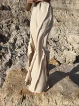 Pantalón largo color beige claro, de tejido ligero con textura, tiro alto y cintura elástica. Presenta detalles deshilachados o con terminación cruda en los bordes verticales de la pierna, creando un efecto drapeado y fluido.