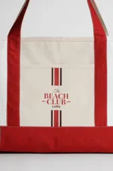 Bolso tipo totebag de lona color crudo con base y asas en color rojo. Estampado frontal con el texto "The Beach Club" en rojo.