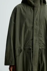 Parka verde militar de tejido técnico ligero, corte oversize, cuello alto con capucha ajustable, manga larga con puños elásticos, cintura ajustable con cordón interno, bolsillos con solapa, bajo asimétrico con abertura trasera ajustable y cierre frontal con cremallera oculta y botones a presión.