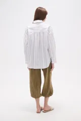 Camisa de algodón blanca de corte holgado, con cuello camisa y manga larga. Presenta un estampado de finas rayas verticales negras y un bolsillo frontal con rayas horizontales.