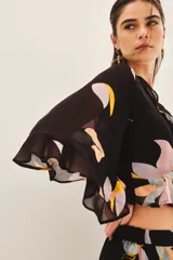 Camisa crop de manga larga con volados en mangas y cintura, confeccionada en viscosa georgette estampada con motivos florales abstractos en tonos pastel sobre fondo negro.