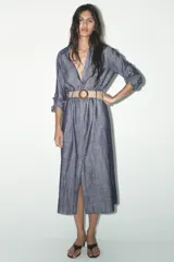 Vestido camisero azul con cinturón de rafia, de cuello solapa y manga tres cuartos con trabilla y botón. Tiene bolsillos de parche en el delantero y cierre frontal con botones.