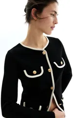 Saco negro de punto con ribetes blancos en cuello, bolsillos y borde inferior. Tiene corte entallado, cuello redondo, cierre frontal con botones dorados y solapas decorativas en el pecho y en la parte inferior.