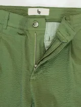 Pantalón de vestir color crema, confeccionado en tejido ligero de algodón con textura seersucker. Tiene corte recto, bolsillos laterales y traseros, cierre con botón y cremallera, y parche con logo de la marca.