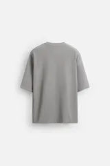 Camiseta gris de corte relaxed fit, cuello redondo y manga corta. Tiene un parche rectangular con texto en el pecho.