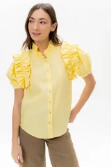 Camisa amarilla de lino con cuello alto con volados y mangas cortas abullonadas con volados. Cierre frontal con botones pequeños.