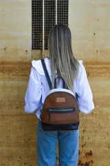 Mochila de cuero color tostado, con base y espalda en negro y tapa superior en blanco. Tiene un bolsillo frontal con cierre y un pequeño aplique de cuero con el logo de la marca.