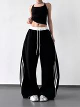 Pantalón deportivo negro de calce extra amplio (wide leg) con cintura elástica blanca y cordón ajustable blanco. Presenta tres tiras verticales blancas a lo largo de las costuras laterales.