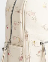 Mochila de cuerina color crema con estampado de flores bordadas, bolsillo frontal con cierre, correas regulables, manija superior, acceso con cierre superior, bolsillos laterales y tag de cuerina.