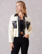 Chaqueta de corte boxy en color crema, confeccionada en tejido de punto suave con detalles en cuero sintético negro en cuello, solapas, cartera frontal y bolsillos. Presenta cierre frontal con botones metálicos plateados, hombros caídos y costuras decorativas horizontales.