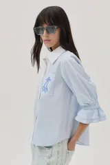 Camisa celeste a rayas verticales blancas, con cuello camisero blanco, mangas 3/4 con volados y bolsillo con bordado de cerezas.