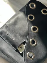 Cartera bandolera de cuero negro con ojalillos metálicos plateados en el frente y correa regulable con hebilla. Tiene un bolsillo exterior y gancho interior para llaves.