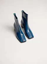 Botas de cuero charol negro con caña media y punta cuadrada. Cuentan con cierre trasero, tirador y bordado contrastante en el empeine. El taco es de madera y la suela de goma.