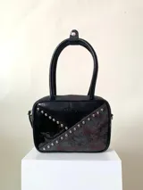 Cartera rígida negra de cuero con tachas plateadas y tres combinaciones de cuero diferentes que aportan textura. Tiene herrajes y cierre metálico plateado marca YKK. Cuenta con un asa fina para llevar al hombro o en la mano y una correa larga extra regulable.