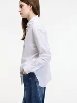 Camisa blanca a rayas verticales finas, con cuello clásico y cierre frontal con botones. Presenta apliques de pedrería en el cuello y en la tapeta de los botones.