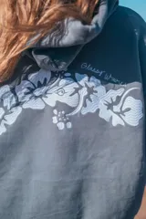 Hoodie gris con capucha y cordones ajustables, de corte holgado y manga larga. Presenta un bordado de flores blancas en la espalda y un pequeño bordado en la manga.