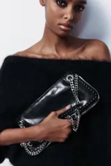 Bolso alargado negro con tachas plateadas en el borde inferior y detalle de cremallera en el corte. Tiene asa de cadena plateada multiposición para usar al hombro o como bandolera. Incluye bolsillo interior con cremallera y cierre principal con cremallera.