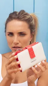 Billetera mediana de símil cuero color blanco hueso con detalles en rojo coral. Posee una presilla de cierre en la parte frontal y cierres de cremallera visibles en el borde superior.