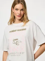 Remera blanca de algodón con estampado frontal de una taza de café, un croissant y la frase "Sunday Morning Coffee Club '87 Fresh Roast All Day".