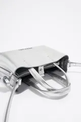 Bolso de mano plateado de piel con estructura rígida y tamaño mini. Tiene logo metálico, acabados metálicos plateados, cierre con cremallera e interior con logo grabado BIMBA Y LOLA.