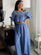 Enterito largo color blanco, con escote Bardot fruncido y mangas cortas abullonadas. La cintura es ajustable con un cinturón ancho de rafia con hebilla redonda. El pantalón es ancho y largo.