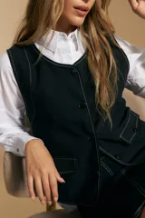 Chaleco negro de vestir con cuello solapa y cierre frontal de botones. Presenta costuras contrastantes blancas que definen el diseño y bolsillos delanteros. Se combina con una camisa blanca de manga larga.