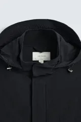 Parka negra de tejido técnico ligero, corte regular, cuello alto con capucha ajustable, manga larga con puños ajustables, cintura ajustable con cordón interno, bolsillos de solapa en la cadera, abertura en la espalda y cierre frontal con cremallera oculta y botones a presión.
