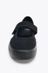 Zapato plano tipo bailarina en tejido color negro. Detalle de pespunte en el cuerpo, tira con adhesivo ajustable, suela engomada y acabado en punta redonda.