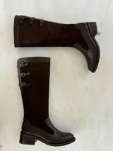 Bota alta de cuero color marrón chocolate, con taco cuadrado de 5 cm y caña alta hasta la rodilla. Presenta tres correas con hebillas en la parte superior de la caña. Fabricada en Colombia.