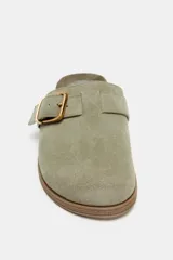 Zueco plano de serraje color verde claro, con tira ajustable y hebilla metálica dorada en la parte delantera. Tiene plantilla aterciopelada, punta redonda y suela de corcho con base de goma.