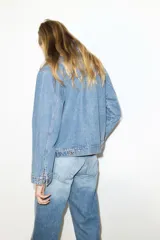 Campera de denim azul de algodón rígido con cuello, cierre delantero y canesú en la espalda. Modelo con puños abotonados, bolsillos delanteros con solapa y bolsillo interior de parche en el pecho. Trabillas ajustables con botones en la parte posterior del bajo.