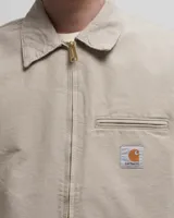 Campera Carhartt modelo Detroit color beige, con cuello camisero, cierre frontal con cremallera, bolsillos laterales y bolsillo en el pecho con logo de la marca.