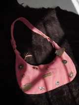 Cartera baguette de gamuza rosa con asa corta y múltiples apliques decorativos: botones, monedas, dijes de metal con forma de sol y caballo, y placa metálica con la marca "Agnes Lenoble".