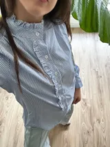 Camisa celeste y blanca a rayas verticales, con cuello con volados, cierre frontal con botones y mangas largas con puños abotonados.