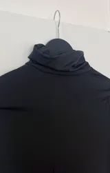 Top negro de manga larga con cuello alto y diseño asimétrico que deja al descubierto un costado del abdomen y cae en un panel largo hasta la rodilla o tobillo.