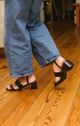 Sandalias plateadas con dos tiras horizontales sobre el pie y una tira vertical que se une a una pulsera con hebilla en el tobillo. La pulsera tiene ojales y tachas combinadas. El taco es cuadrado y de 5 cm.